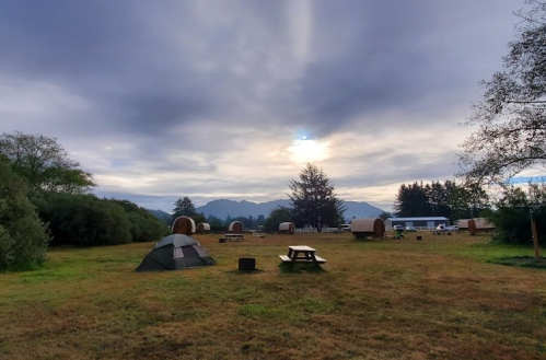 A serene campsite with a tent, picnic table, and wooden cabins under a cloudy sky and distant mountains.