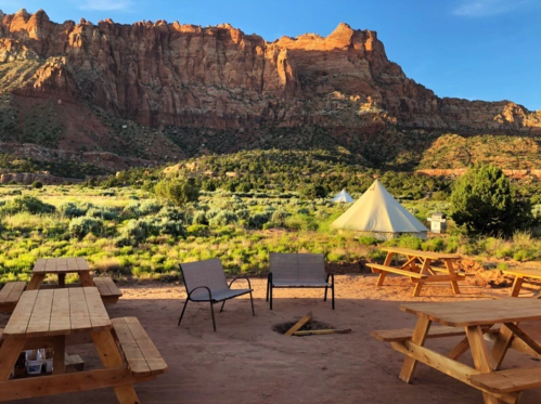 A scenic campsite with wooden tables and chairs, surrounded by lush greenery and towering red rock formations.