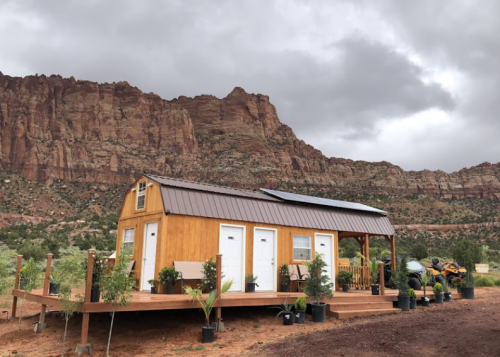 A small wooden house with a porch, surrounded by plants, set against a backdrop of rocky cliffs and cloudy skies.