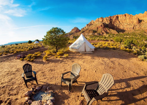 A glamping site with a tent and two chairs, surrounded by desert landscape and mountains under a clear blue sky.