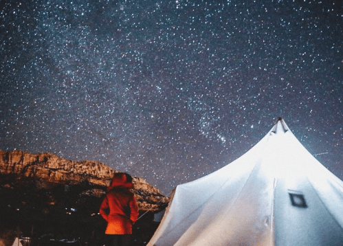 A person in a red jacket stands near a tent, gazing up at a starry night sky filled with stars.