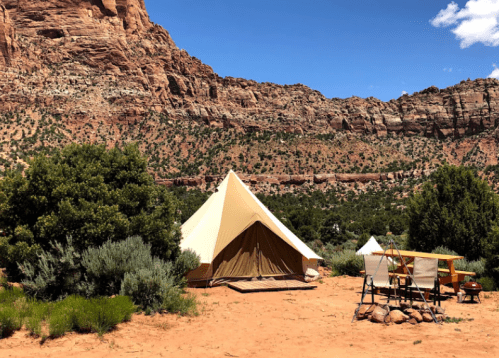 A glamping tent set up in a desert landscape, surrounded by shrubs and rocky cliffs under a clear blue sky.