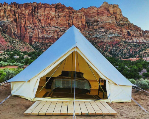 A spacious tent with a bed inside, set against a backdrop of colorful rock formations and greenery.