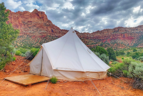 A white tent set against red rock formations and a cloudy sky, surrounded by greenery in a desert landscape.