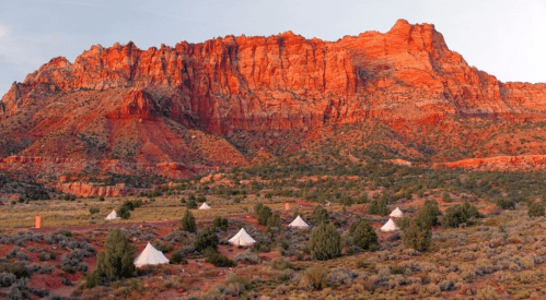 A desert landscape with white tents in the foreground and vibrant red rock formations in the background at sunset.