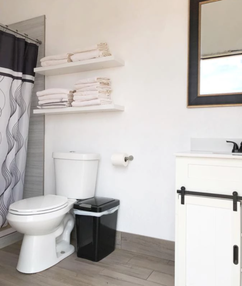 A clean bathroom featuring a toilet, sink, mirror, shelves with towels, and a trash can.