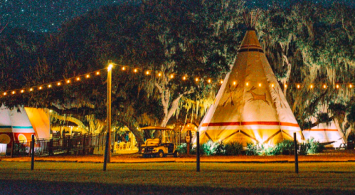 A serene night scene of illuminated teepees surrounded by trees and string lights under a starry sky.