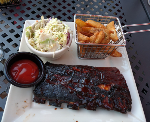 A plate featuring BBQ ribs, coleslaw, and seasoned potato wedges with a side of ketchup.