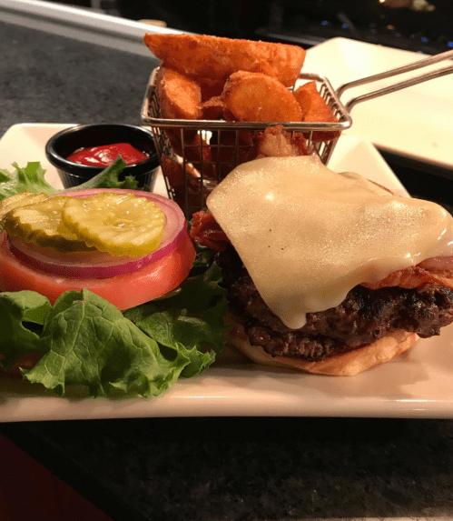 A juicy burger with melted cheese, lettuce, tomato, pickles, and bacon, served with crispy potato wedges and ketchup.