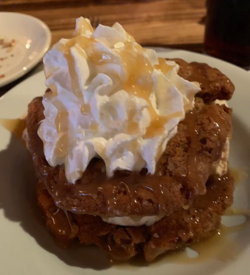A dessert featuring two chocolate chip cookies layered with ice cream, topped with whipped cream and caramel sauce.