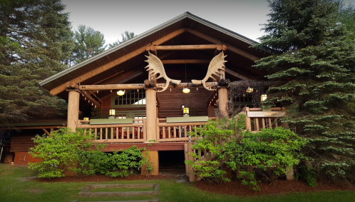A rustic wooden cabin with large moose antlers on the front, surrounded by trees and greenery.