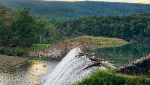 A serene waterfall cascades over a stone dam, surrounded by lush greenery and mountains in the background.