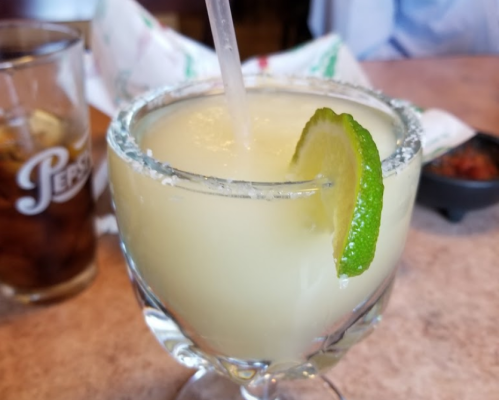A close-up of a margarita in a glass with a salted rim and a lime wedge, next to a glass of cola.