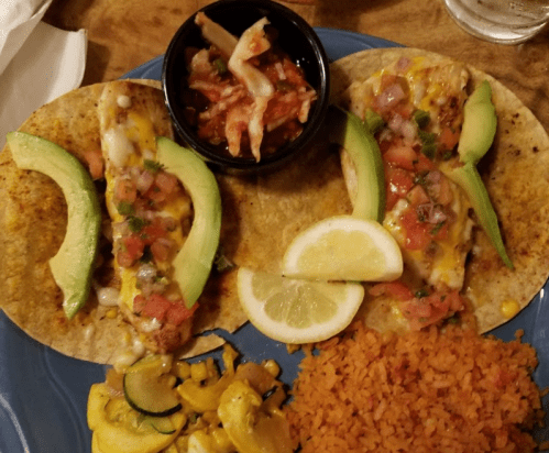 Two tacos topped with avocado, salsa, and cheese, served with rice and corn on a blue plate.