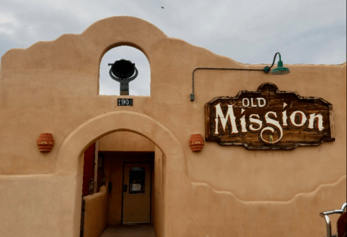Exterior of a building with a sign reading "Old Mission," featuring adobe-style architecture and a bell.