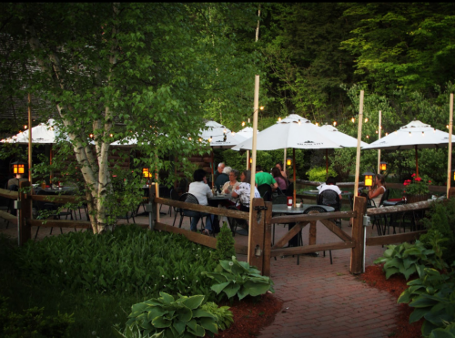 Outdoor dining area with tables under umbrellas, surrounded by greenery and soft lighting, creating a cozy atmosphere.