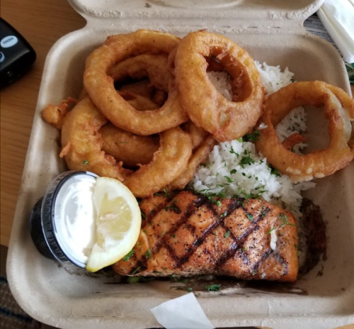 A takeout box with grilled salmon, rice, onion rings, a lemon wedge, and a small container of sauce.