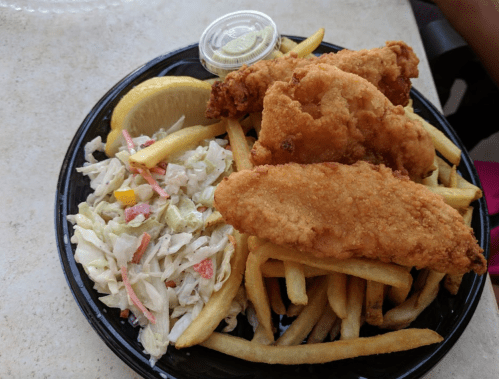 A plate of fried fish fillets served with French fries, coleslaw, a lemon wedge, and a small container of sauce.
