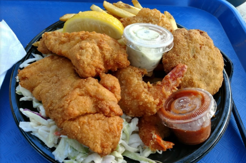 A plate of fried fish, shrimp, hushpuppies, fries, and lemon, served with dipping sauces on a blue tray.