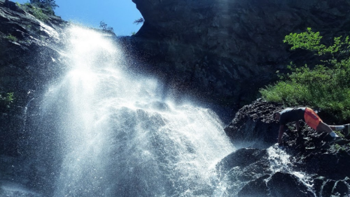 A person climbs near a waterfall, with sunlight shining through the mist and lush greenery surrounding the rocky area.