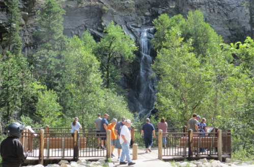 A group of people stands on a viewing platform near a waterfall surrounded by lush greenery and rocky cliffs.