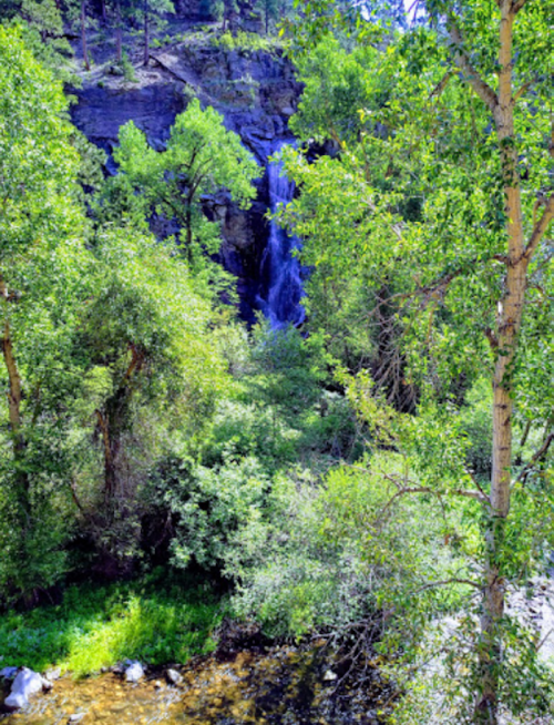 A serene waterfall cascades down rocky cliffs, surrounded by lush green trees and a tranquil stream below.