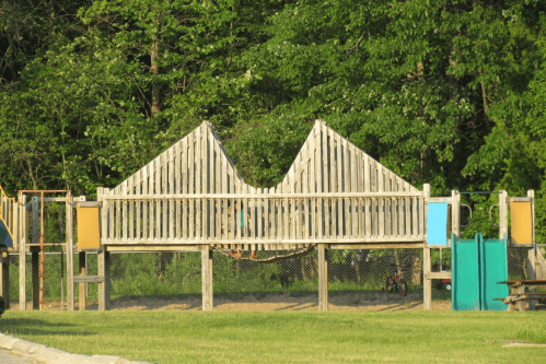 A wooden playground structure with a unique design, surrounded by greenery and a grassy area.