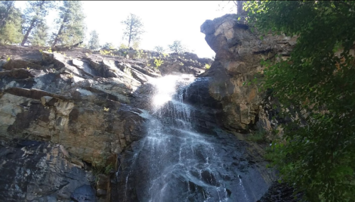 A cascading waterfall flows down rocky cliffs, surrounded by trees and sunlight filtering through the foliage.