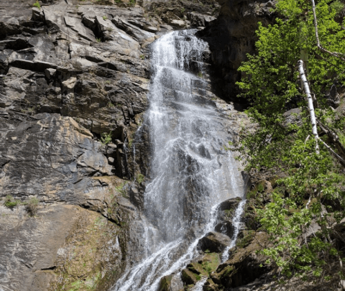 A cascading waterfall flows down rocky cliffs, surrounded by lush greenery and rugged terrain.