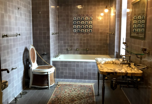A vintage bathroom featuring a bathtub, marble sink, patterned tiles, and a woven chair on a patterned rug.