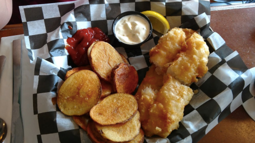 A plate of crispy fried fish fillets with potato chips, ketchup, tartar sauce, and a lemon wedge on a checkered paper.