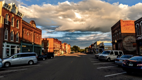 A quiet street in a small town, lined with historic brick buildings and parked cars under a cloudy sky.