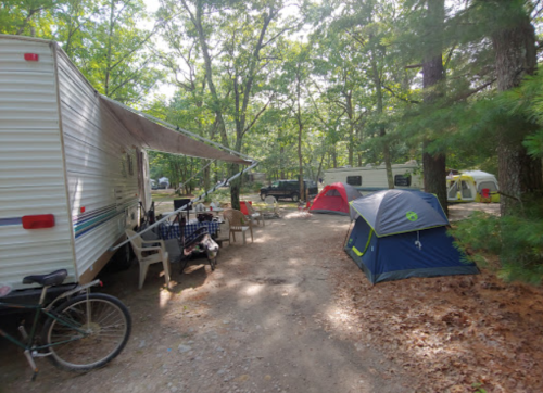 A campsite with a trailer, tents, picnic table, and bike surrounded by trees and fallen leaves.