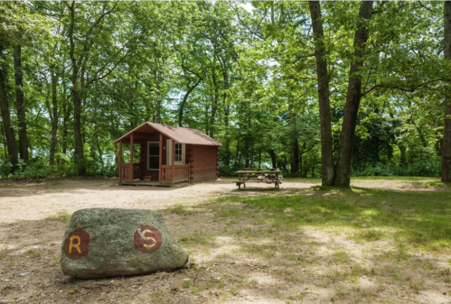 A small wooden cabin surrounded by trees, with a picnic table nearby and a marked rock in the foreground.