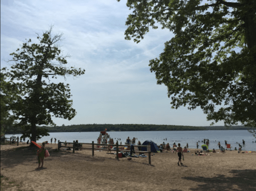 A sandy beach scene with people swimming, sunbathing, and a lifeguard tower under a clear sky and trees.
