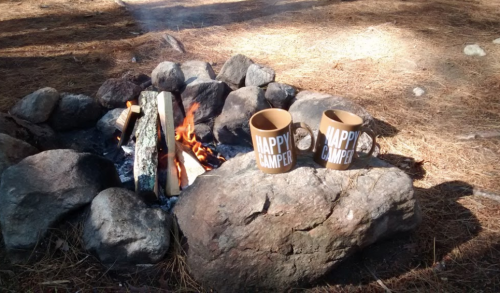 Two "Happy Camper" mugs sit on rocks beside a campfire, surrounded by pine needles and stones.