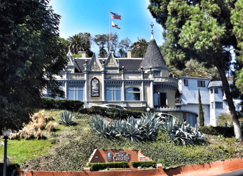 A grand, castle-like building with turrets, surrounded by greenery and featuring the sign for "The Magic Castle."