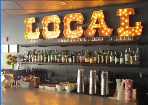 A bar with a "LOCAL" marquee sign, shelves of liquor bottles, and a display of fruits and garnishes.