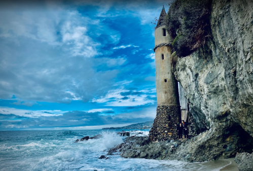 A tall, weathered tower stands on a rocky beach, with waves crashing against the shore under a cloudy sky.