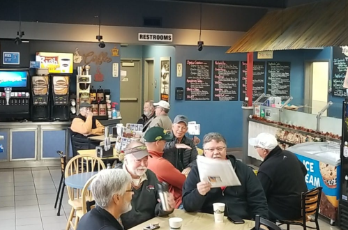 A cozy café scene with several men chatting and reading newspapers at tables, with a menu board in the background.