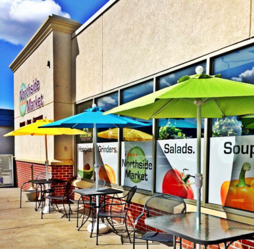 Outdoor seating area with colorful umbrellas at Northside Market, featuring signs for hoagies, grinders, salads, and soups.