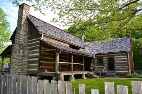 A historic log cabin with a stone chimney, surrounded by greenery and a wooden fence.