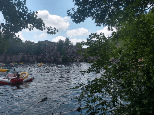 A serene lake surrounded by trees, with people floating on colorful inflatables under a partly cloudy sky.