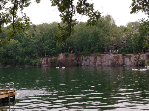 A serene lake surrounded by lush greenery, with people on a rocky ledge and a wooden dock in the foreground.