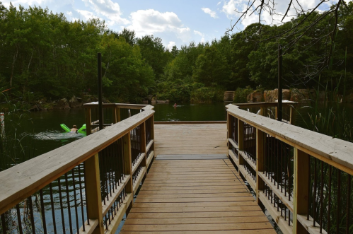 A wooden dock leads to a serene lake surrounded by trees, with people swimming in the water.
