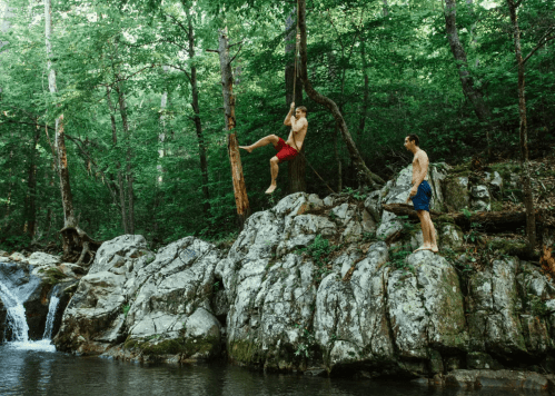 Two men in swim trunks near a rocky riverbank, one climbing a tree while the other watches. Lush green forest surrounds them.