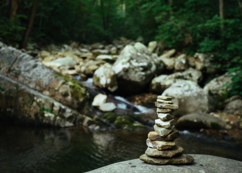 A stack of smooth stones balanced on a rock near a serene stream surrounded by lush greenery.