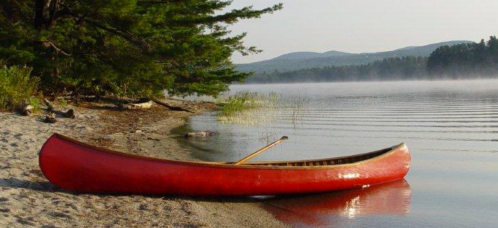 A red canoe rests on a sandy shore by a calm lake, surrounded by trees and misty mountains in the background.