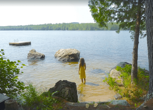 A person in a yellow dress stands in shallow water by a lake, surrounded by rocks and trees.