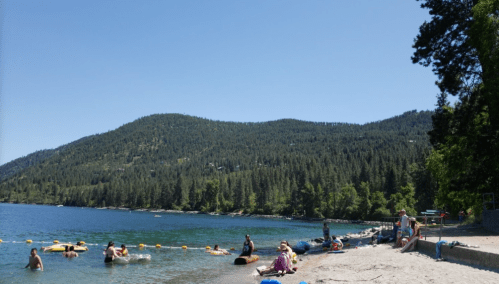 A sunny beach scene with people relaxing by the water, surrounded by trees and mountains in the background.